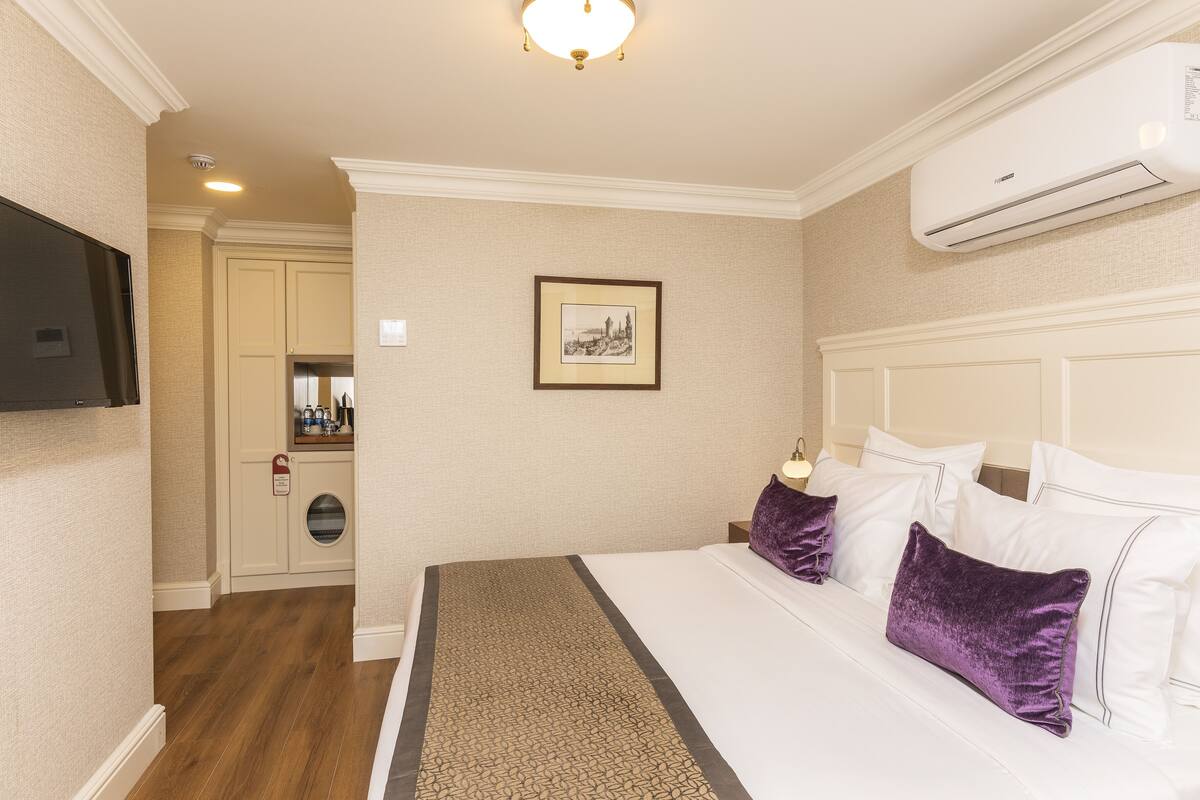 A hotel room with a bed, a wall-mounted air conditioner, a framed picture, and a small kitchenette.