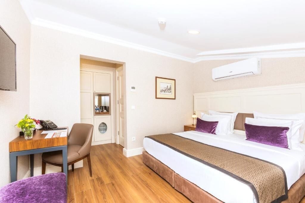 A hotel room with a bed, a desk with a chair, a small table with a plant, and an air conditioning unit on the wall.
