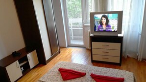 Una camera d'albergo con un letto, una televisione, un pavimento in legno, una finestra con tende e un tavolino con un telefono.
