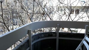 Una panca curva con struttura in metallo e seduta bianca, situata in un’area esterna parzialmente riparata e con vista su alberi spogli e edifici sullo sfondo.