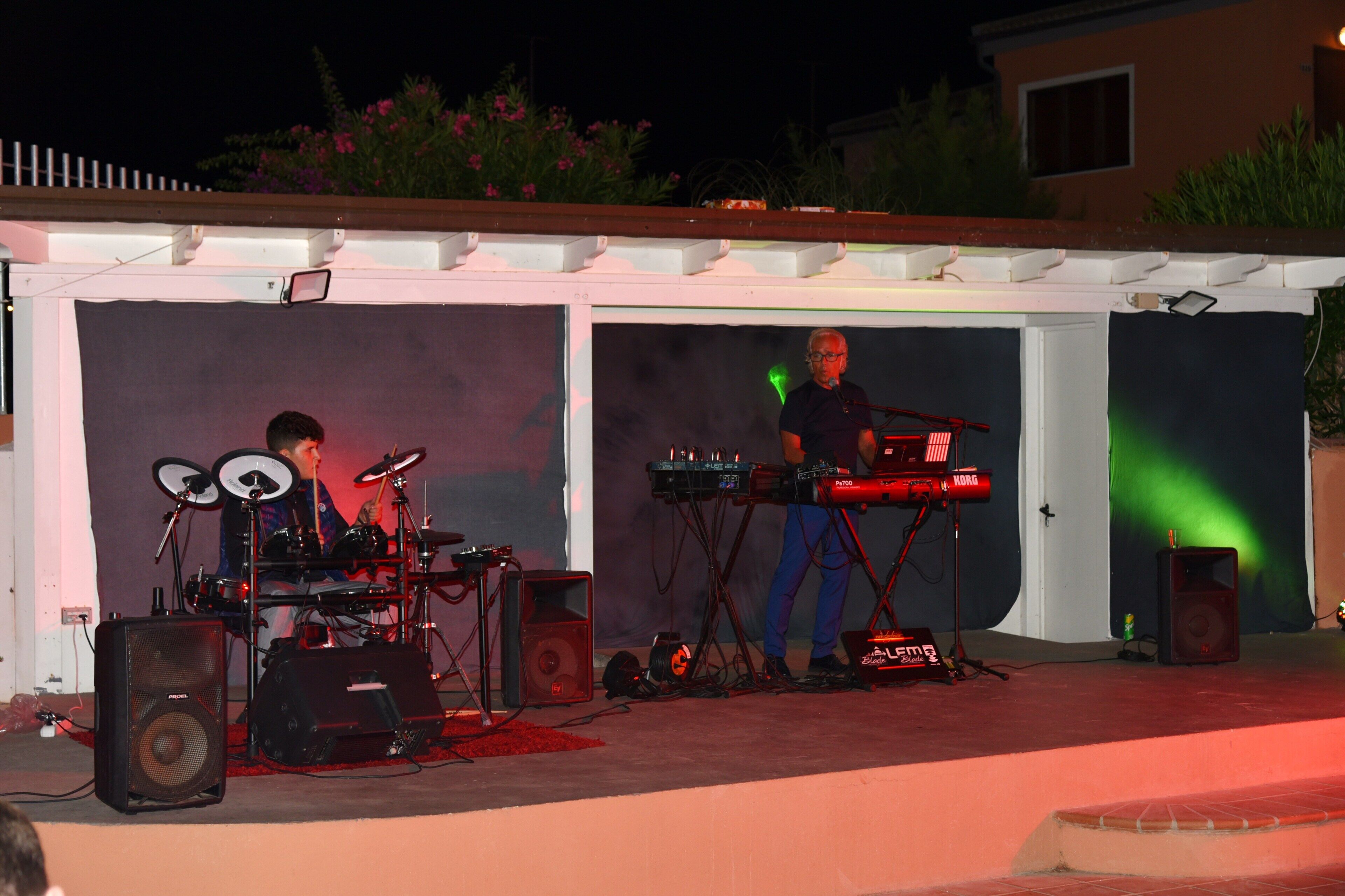 Un escenario al aire libre con equipo musical, incluyendo una batería y un teclado, bajo una estructura cubierta con techo blanco.