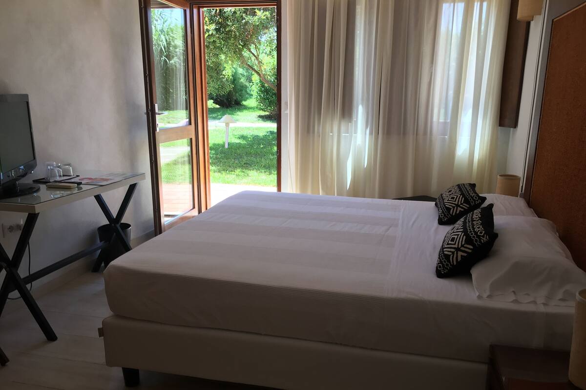 A hotel room with a bed, a desk, and a view of a garden through a glass door.