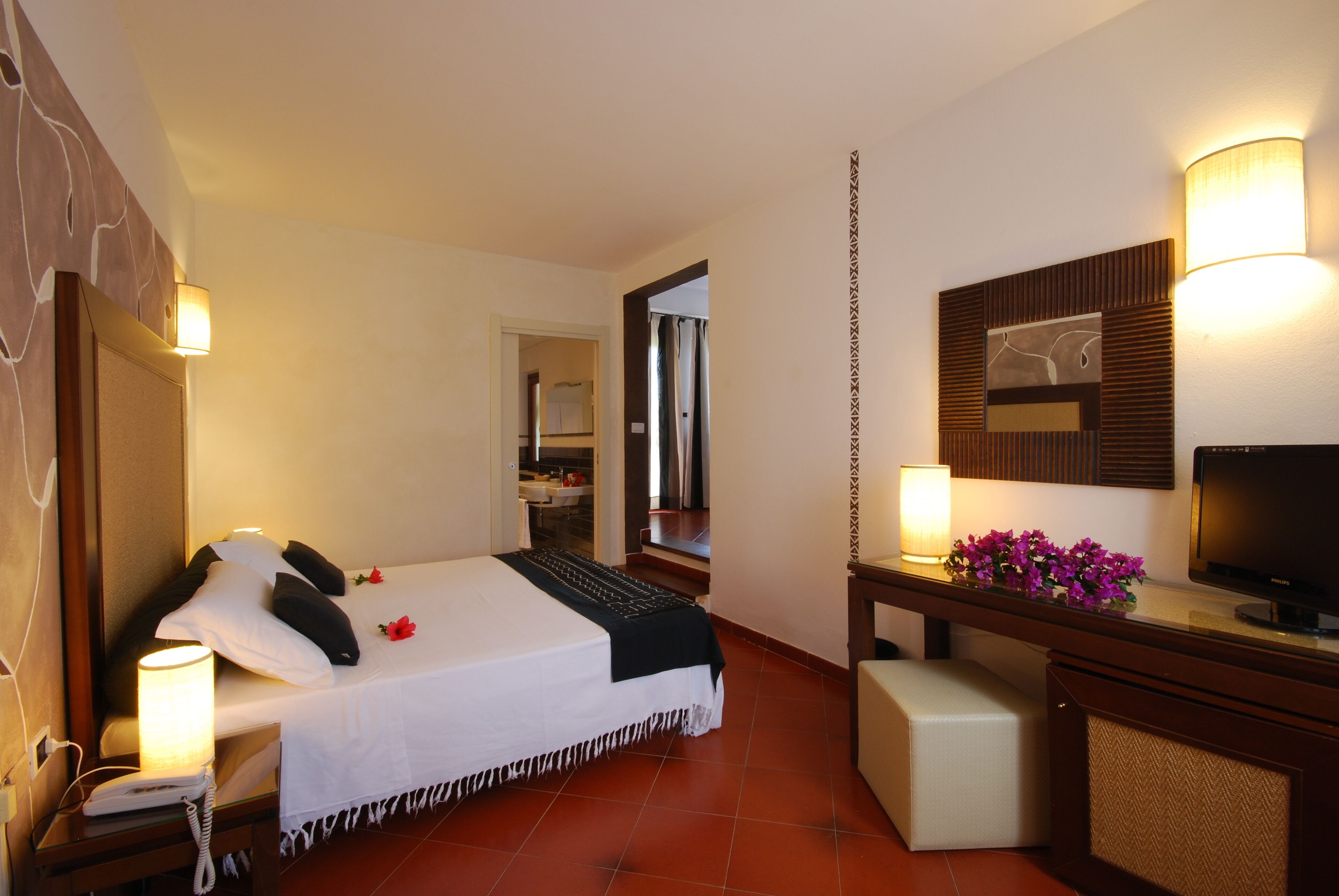 A hotel room with a bed, bedside lamps, a desk with a television, and a bathroom visible through an open door.