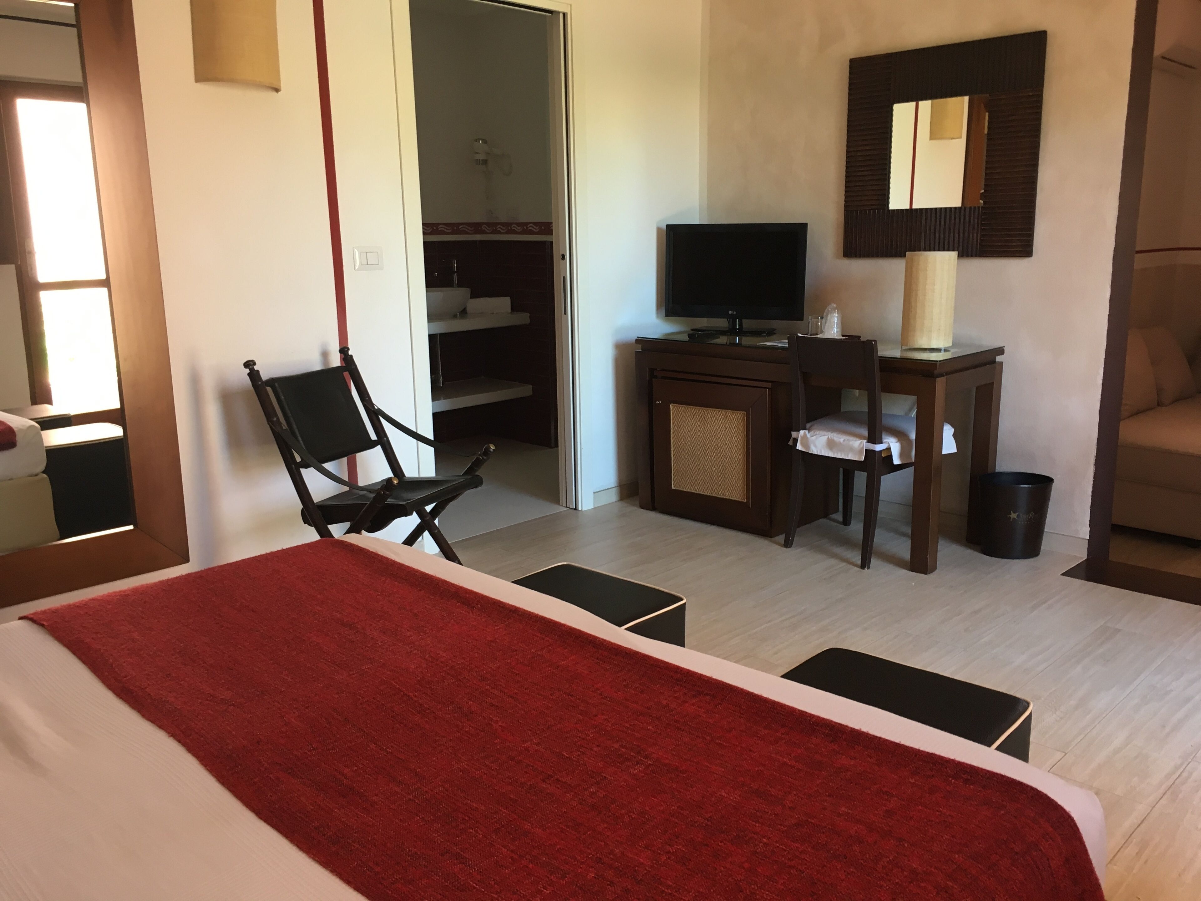 A hotel room with a bed, a desk with a computer, a chair, a TV, and a bathroom visible through an open door.