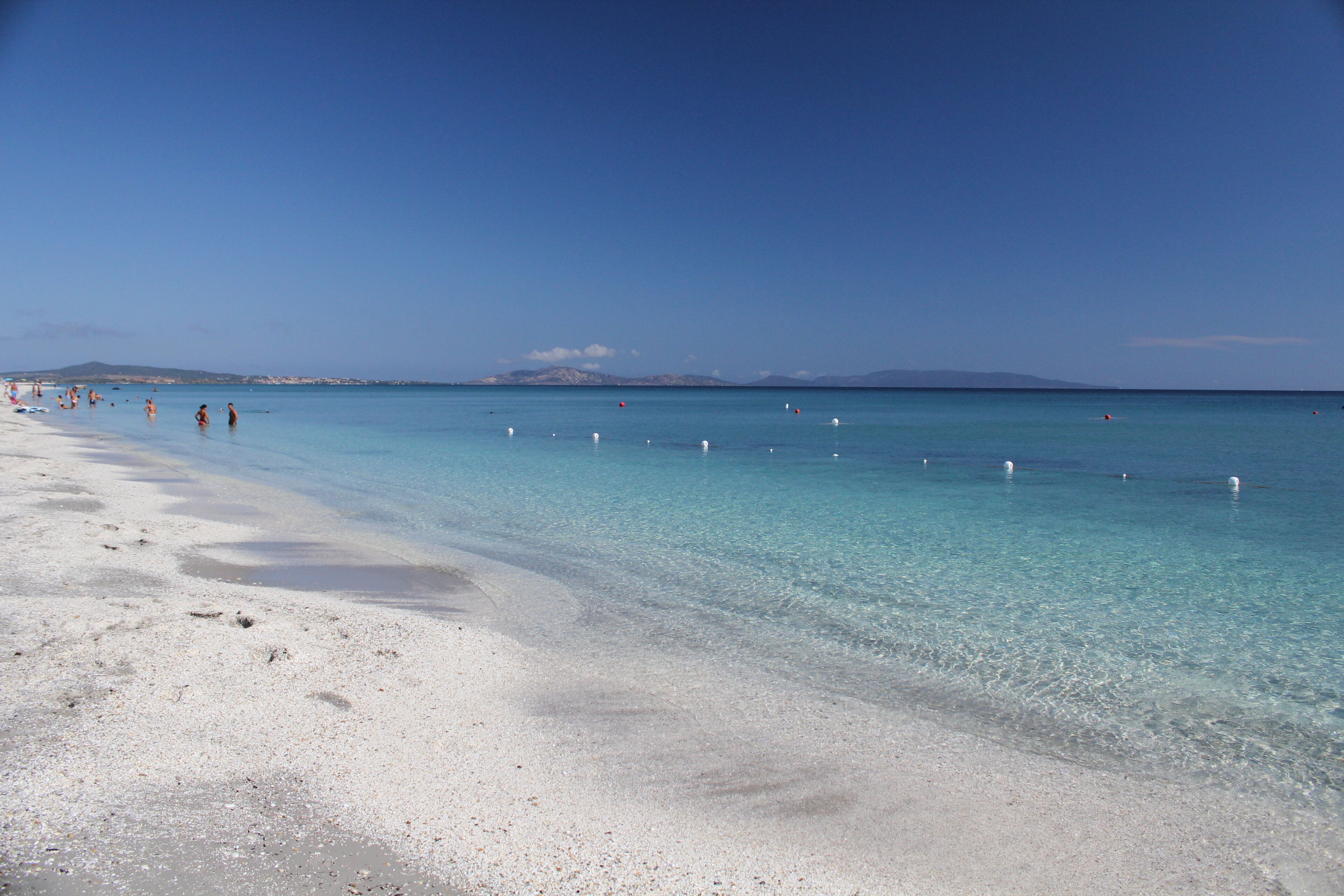 A beach with clear turquoise water, white sand, and a few people in the distance.