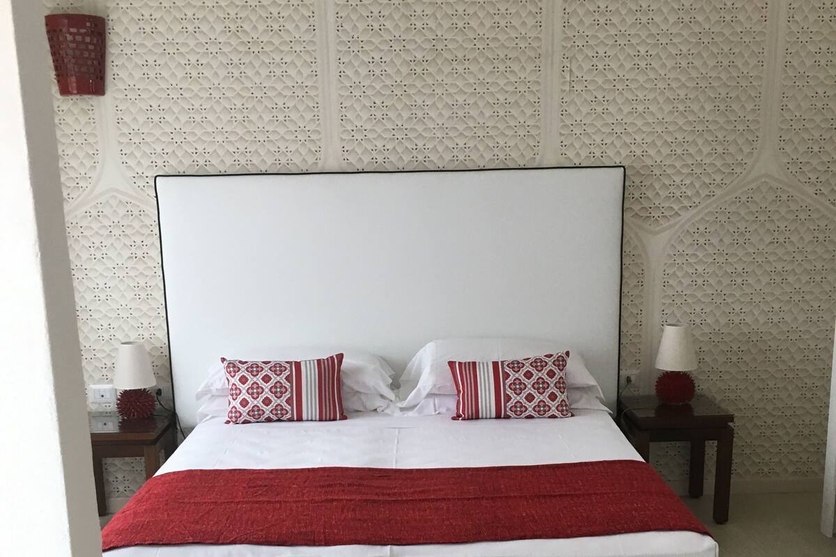 A bed with a white headboard, two pillows with red and white patterns, and a red blanket. There are two bedside tables with lamps. The room has a patterned wallpaper and a light-colored wooden floor.