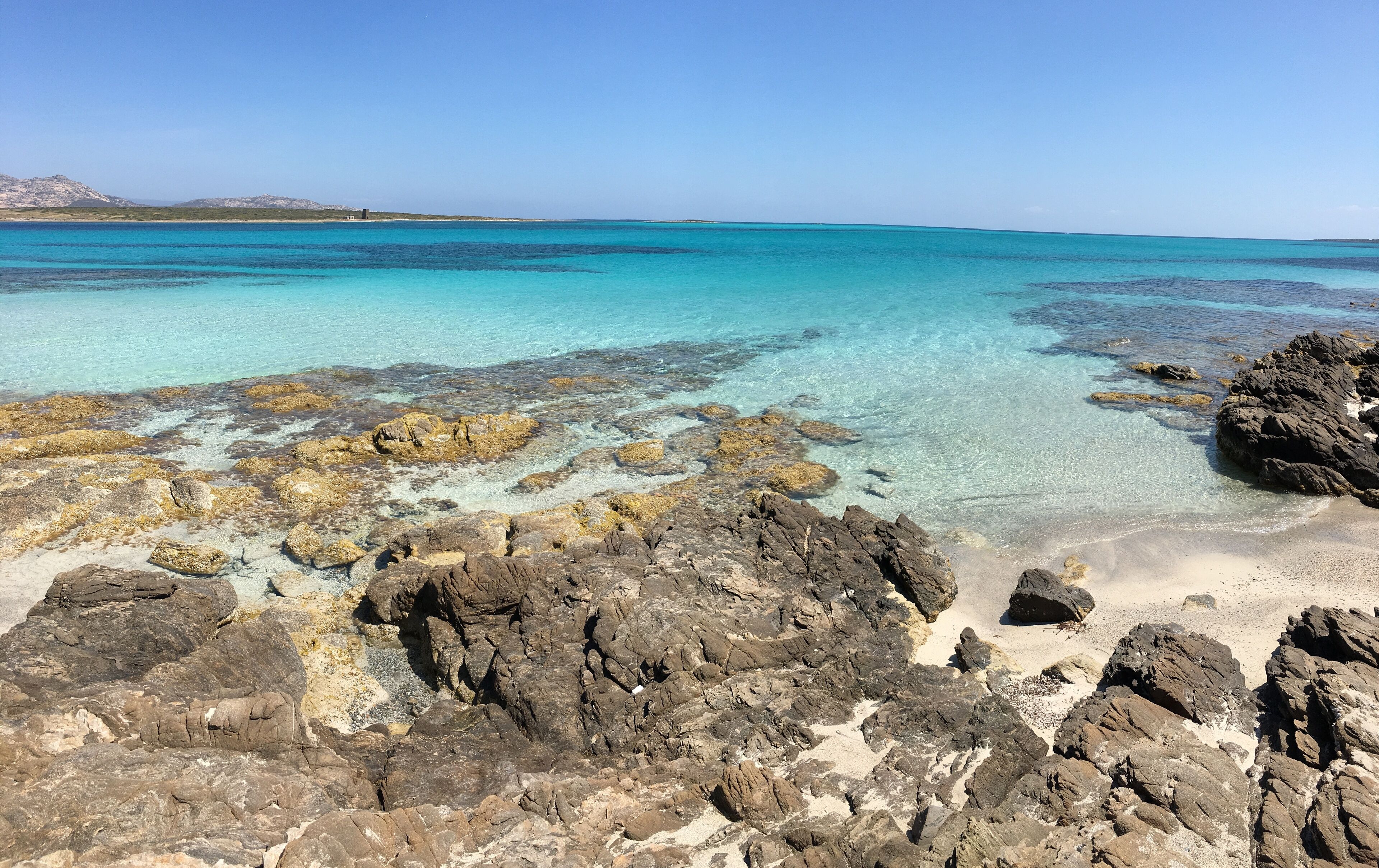 Een rotsachtige kustlijn met helder turquoise water en bergketens in de verte.