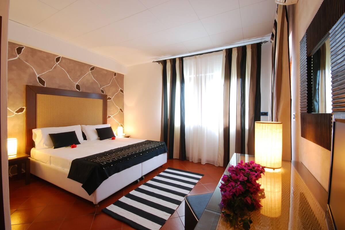 A hotel room with a large bed, a bedside table with a lamp, a striped rug, and a flower arrangement.