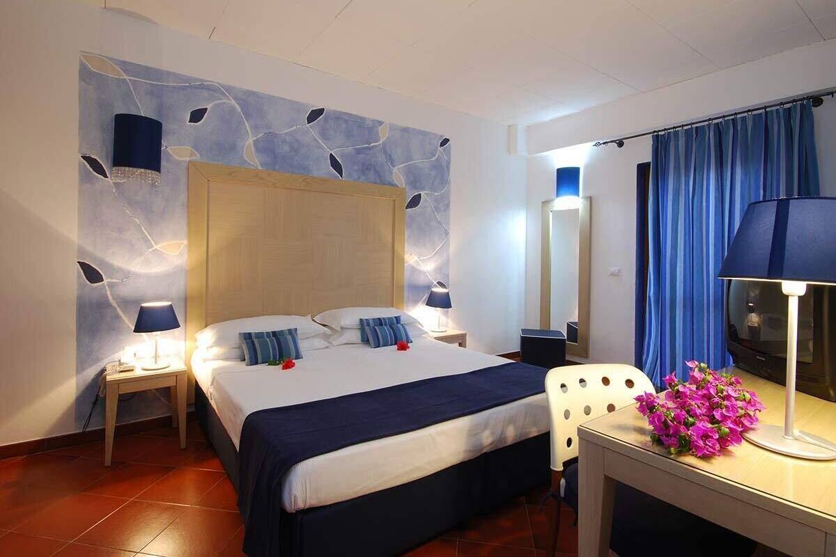 A hotel room with a large bed, bedside lamps, a desk with a chair, a TV, and a decorative wall with leaf patterns.