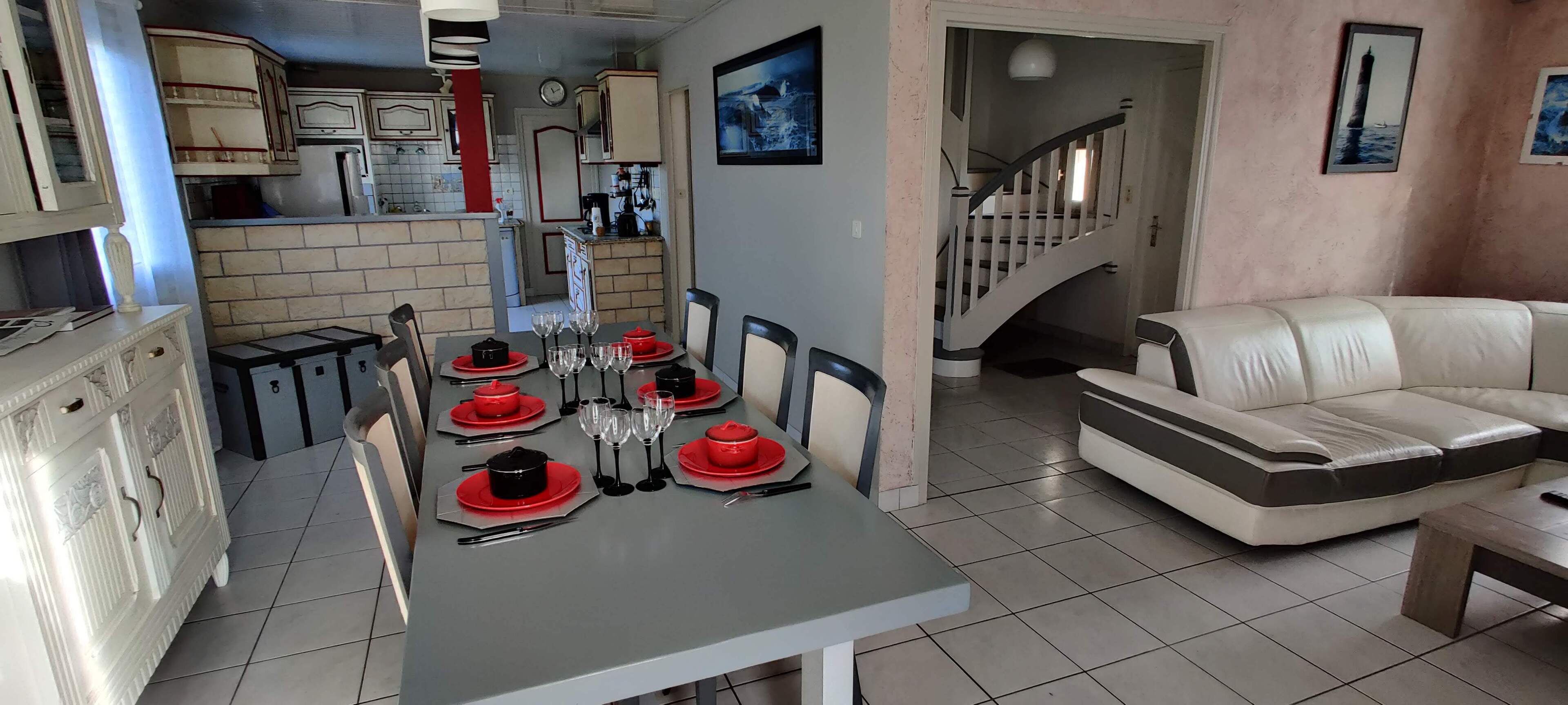 Ein Essbereich mit einem Tisch für vier Personen, eine Küche im Hintergrund und eine Treppe, die zu einer weiteren Etage führt.