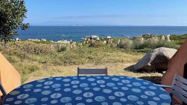 Mesa con mantel azul y silla, con vista a un paisaje costero con rocas y vegetación.