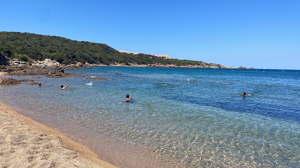 Una playa con agua cristalina, costa rocosa y un paisaje montañoso al fondo.