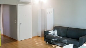 Een woonkamer met een donkere bank, een witte tafel en een witte stoel. Er is een witte deur en een witte kast met haken aan de muur.