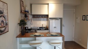 Una cocina compacta con armarios blancos, un refrigerador y un comedor con dos sillas y una mesa blancos.
