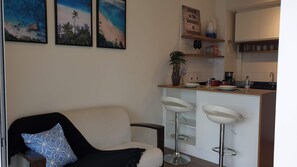 Sala de estar con un sofá y dos taburetes blancos, una zona de cocina con mostrador, y una pared decorada con tres cuadros enmarcados.