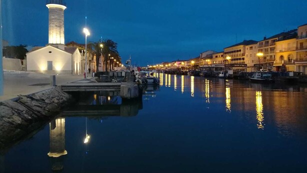 Escena costera con un faro destacado, agua tranquila que refleja luces y edificios a lo largo de la orilla.