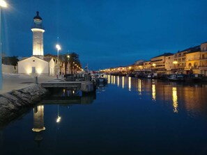 A waterfront scene with a prominent lighthouse, calm water reflecting lights, and buildings along the shore.