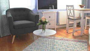 A room with a grey armchair, a small round table with a vase of flowers, a white desk, and a television on a stand. - Nice apartment lilac central (Bremen)