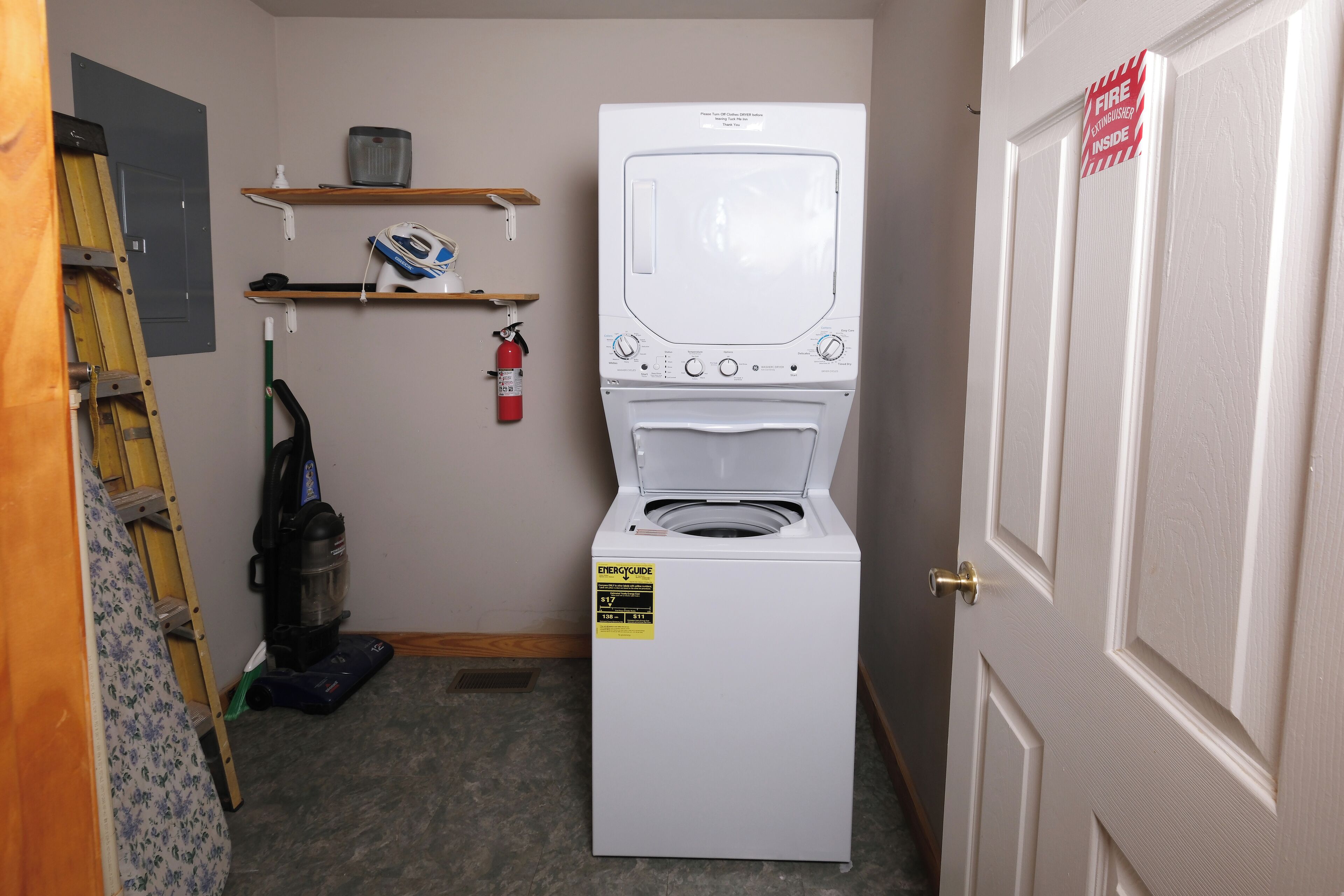 A laundry room with a front-loading washer and dryer, a fire extinguisher, and a shelf with cleaning supplies.