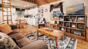 A cozy living room with a sofa, coffee table, and a bookshelf. A kitchen with white cabinets and a dining area with a table and chairs are visible. - Couples, Honeymoon, Anniversary Cabin, convenient to Gatlinburg, National Park (Gatlinburg)