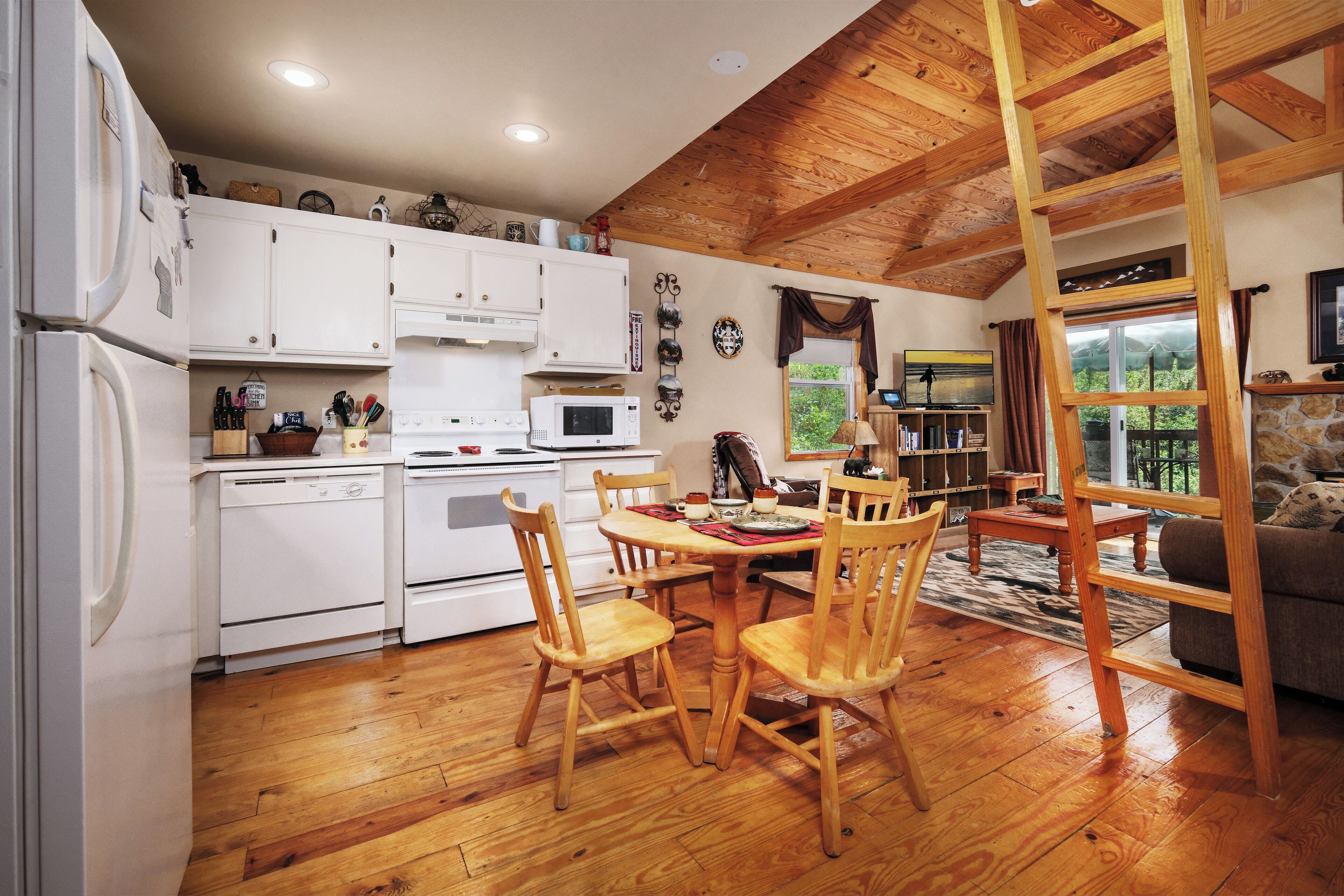A kitchen with white cabinets, a wooden dining table with chairs, a wooden ladder, and a stone fireplace.