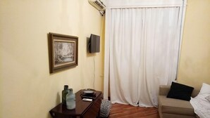 Een kamer met een houten vloer, een bureau met stoel, een bed met kussen, een ingelijste foto aan de muur en een raam met witte gordijnen.