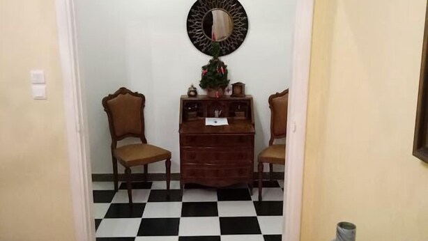 Een gang met een geruit vloerkleed, een klein houten dressoir, twee stoelen en een spiegel aan de muur.
