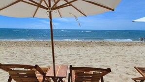 A beach with wooden chairs and tables under a large umbrella, overlooking the ocean. - Carambola Hotel (Porto Seguro)