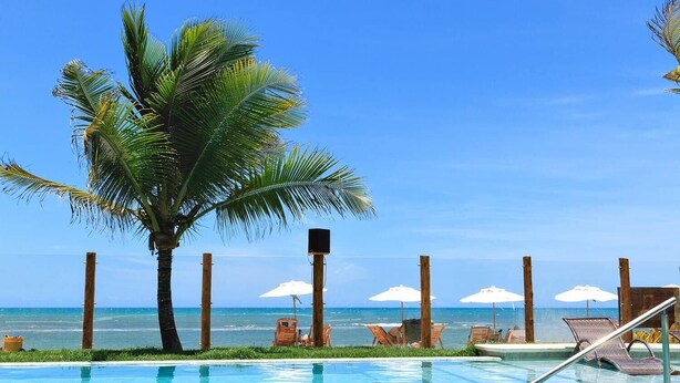 A pool area with lounge chairs and umbrellas, palm trees, and a view of the ocean. - Carambola Hotel (Porto Seguro)
