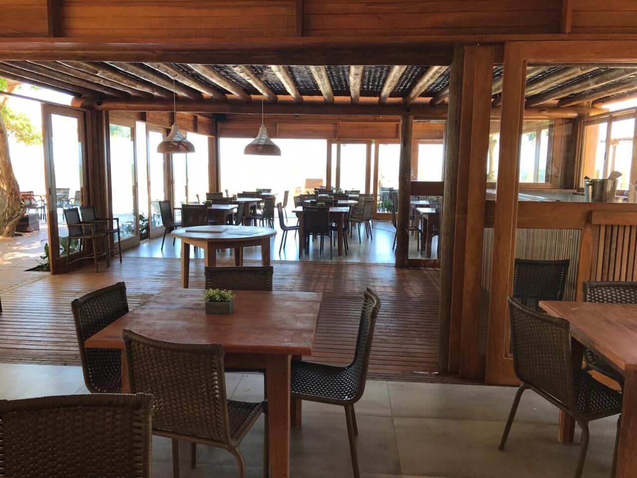 A spacious outdoor dining area with wooden tables and chairs, large windows, and a wooden ceiling structure.