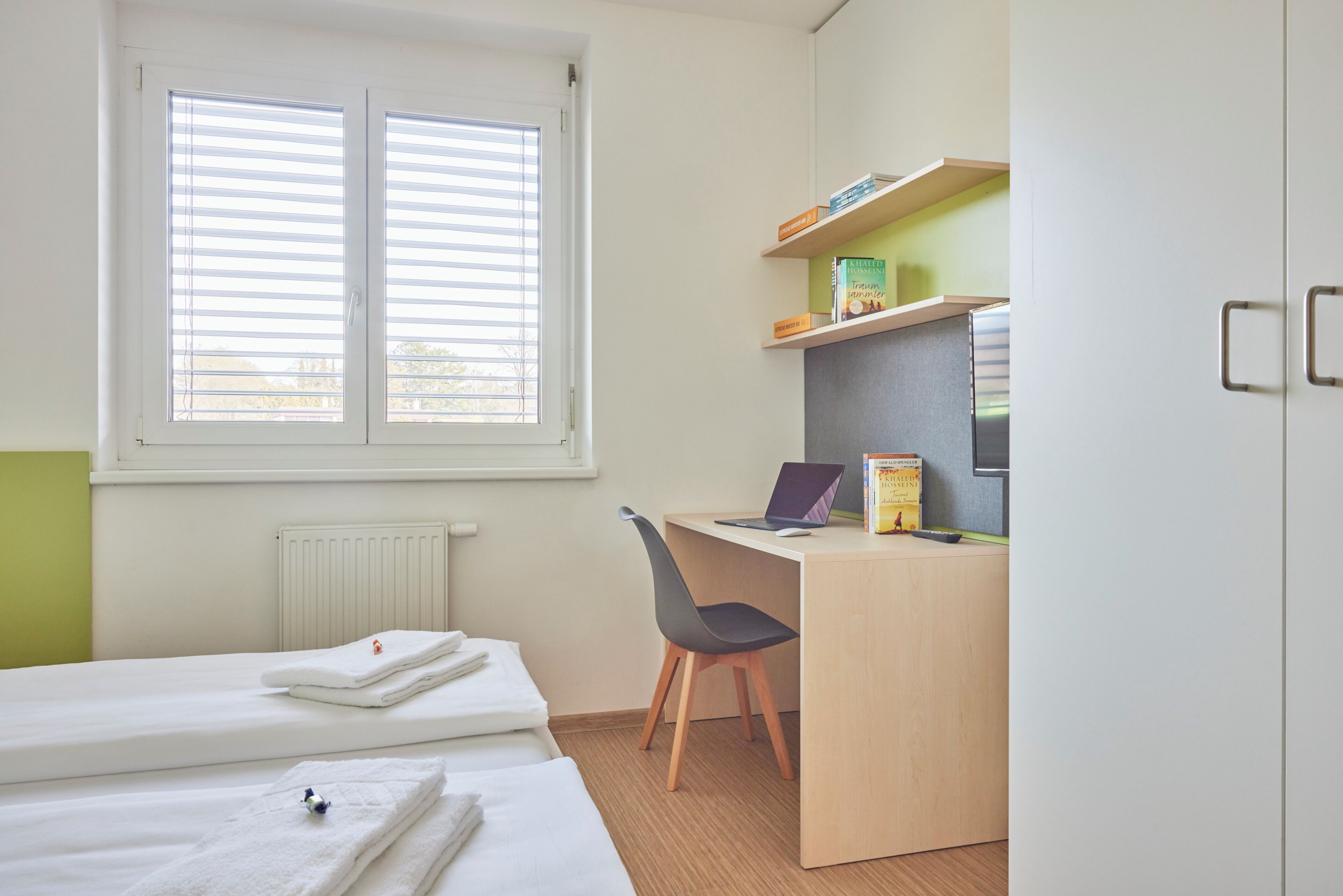 Un dormitorio con cama, escritorio, ordenador portátil, silla y ventana con persianas.