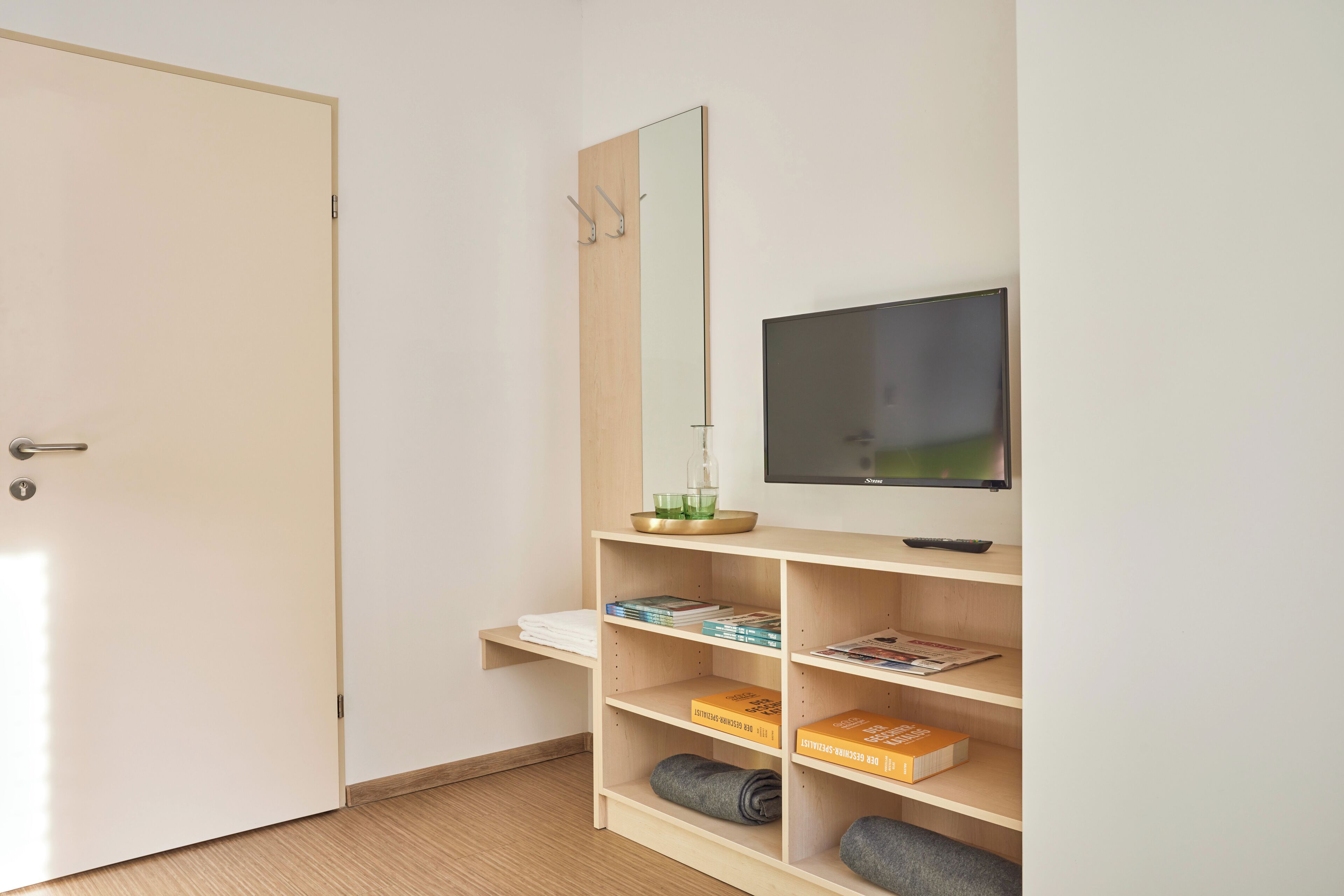 A room with a door, a mirror, a television, and shelves with books and a rolled-up blanket.