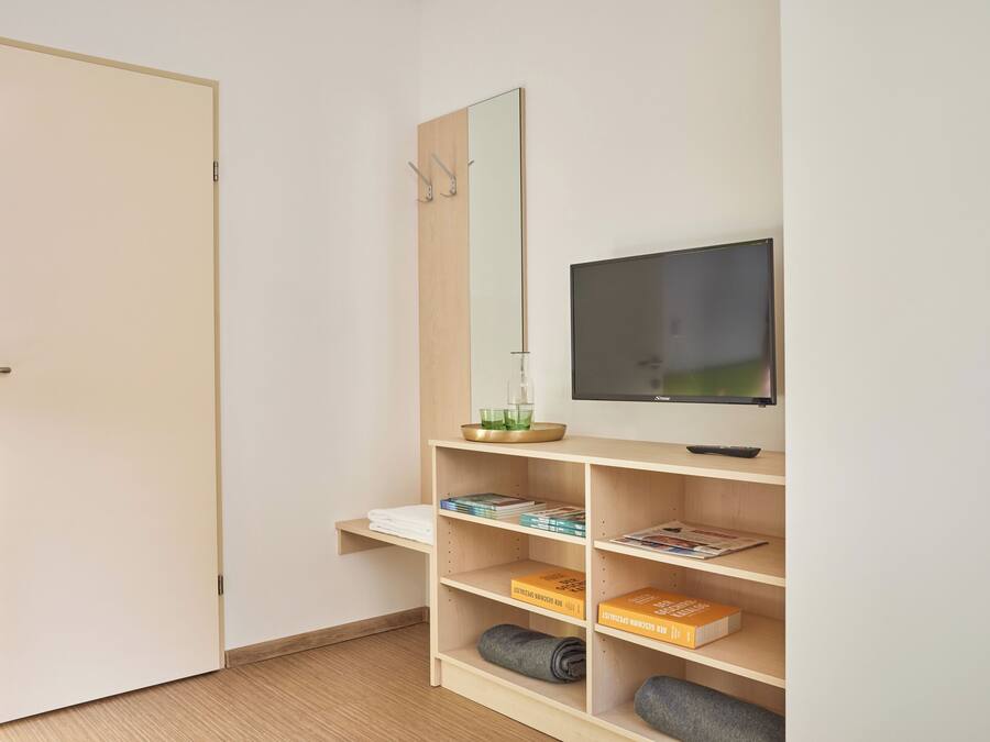 A room with a door, a mirror, a television, and shelves with books and a rolled-up blanket.
