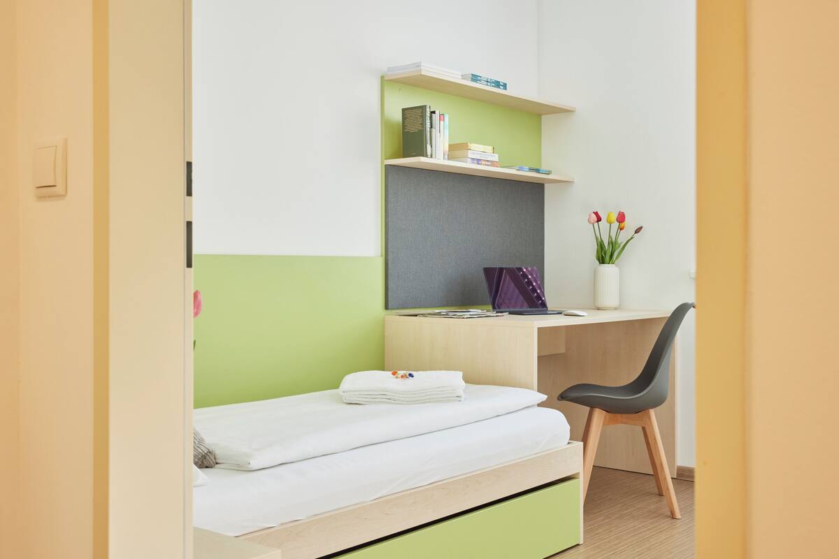 A room with a bed, a desk with a chair, a shelf with books, and a vase with flowers.