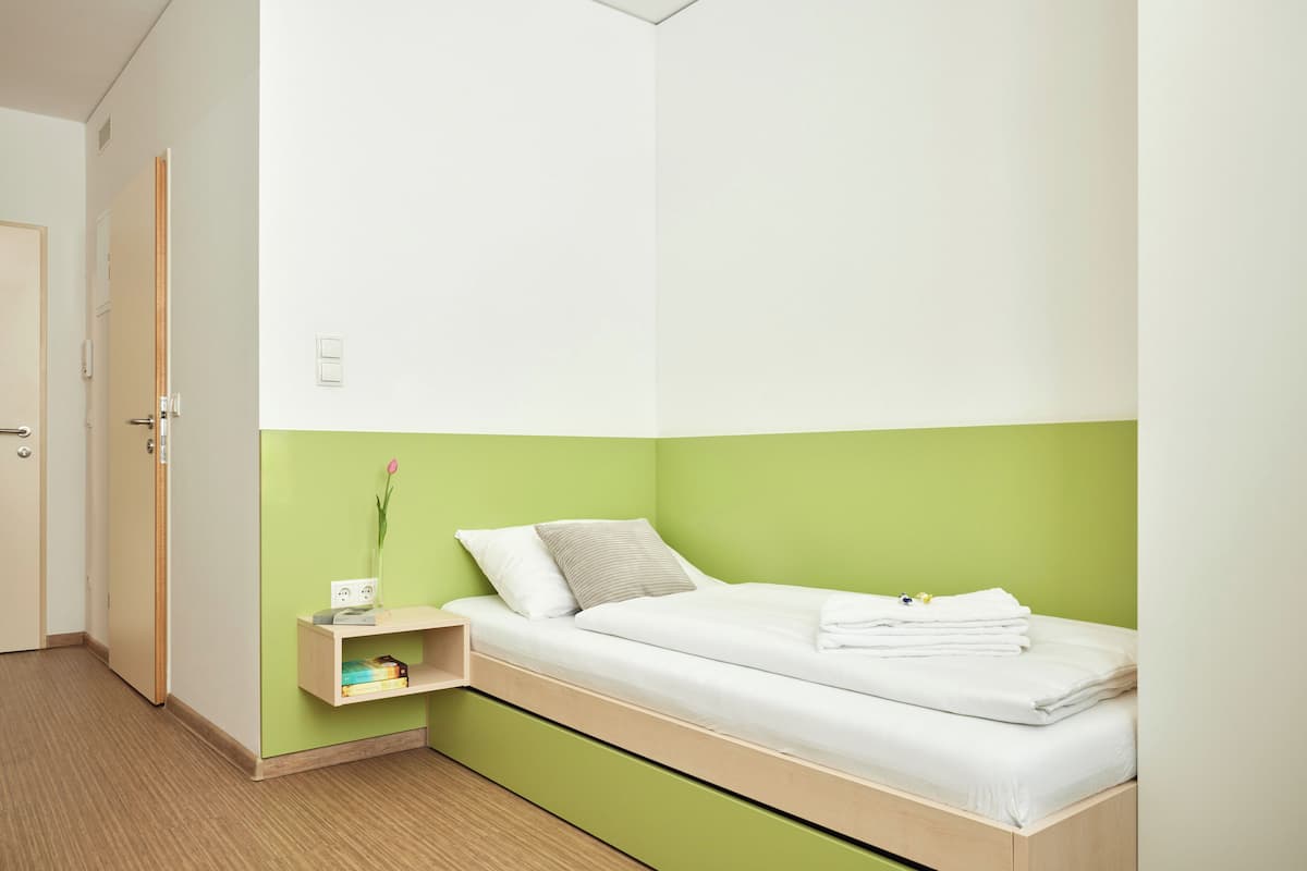 Habitación con cama individual, pared de acento verde, mesita de noche con un florero y una puerta a la izquierda.