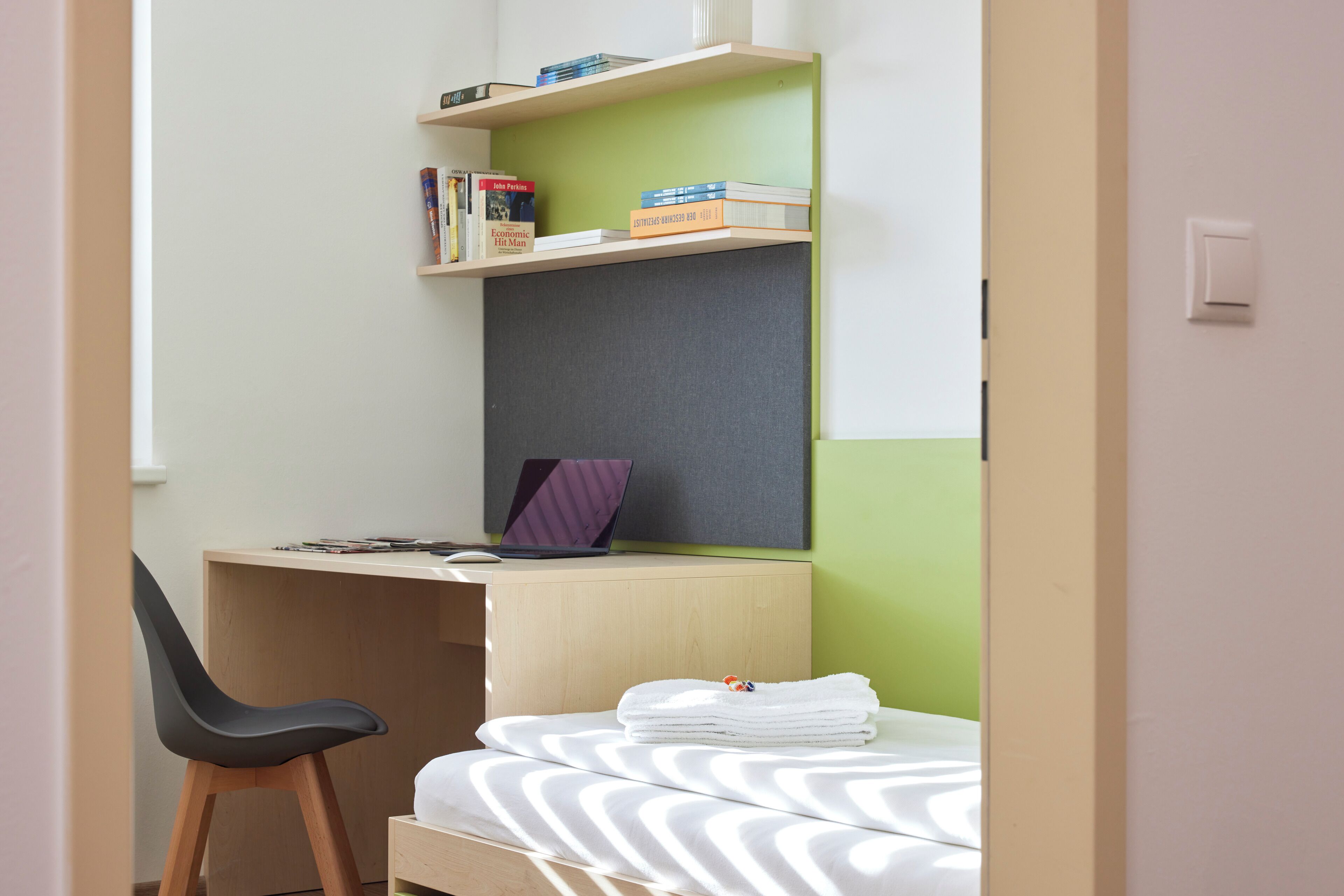 Una habitación pequeña y ordenada, con escritorio, silla y cama, que incluye una computadora portátil, libros y una toalla doblada con cuidado.