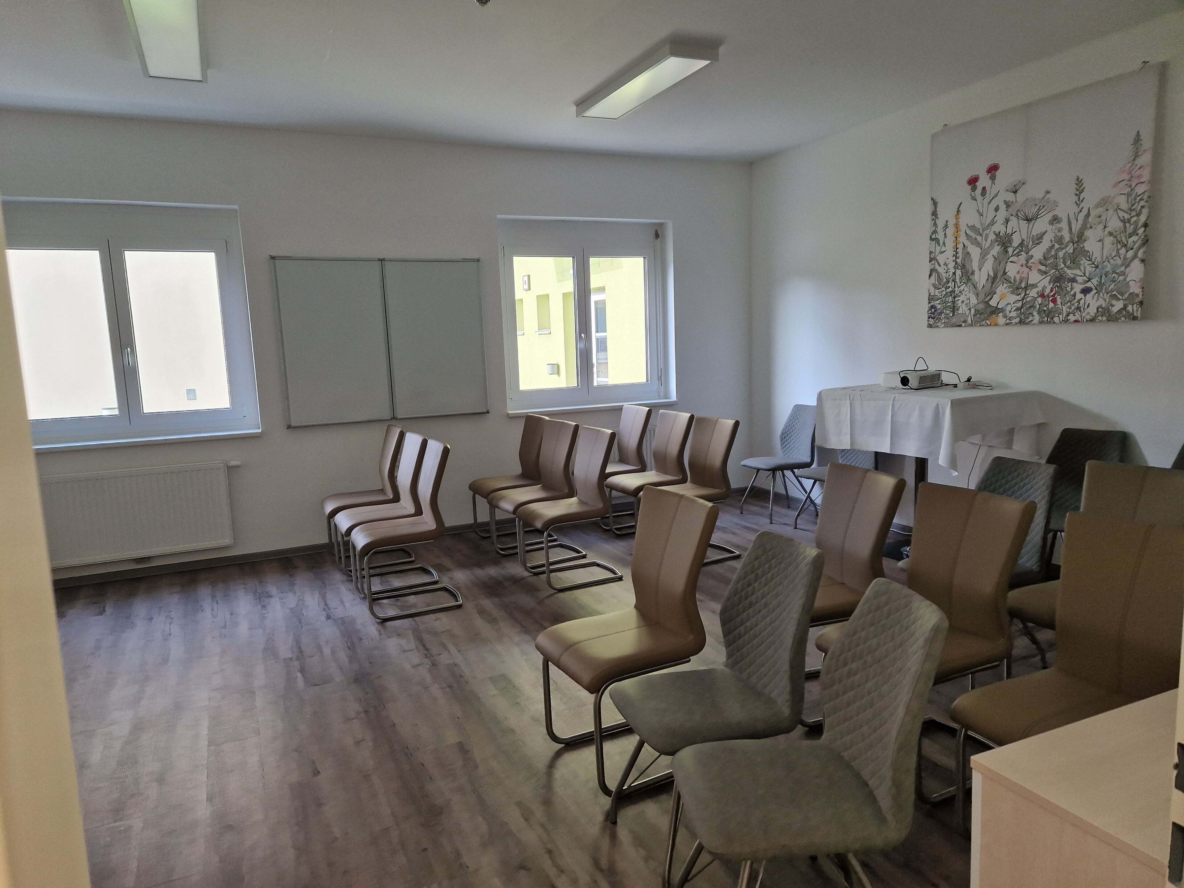 A room with multiple chairs arranged in rows, a table with a white tablecloth, and a painting on the wall.