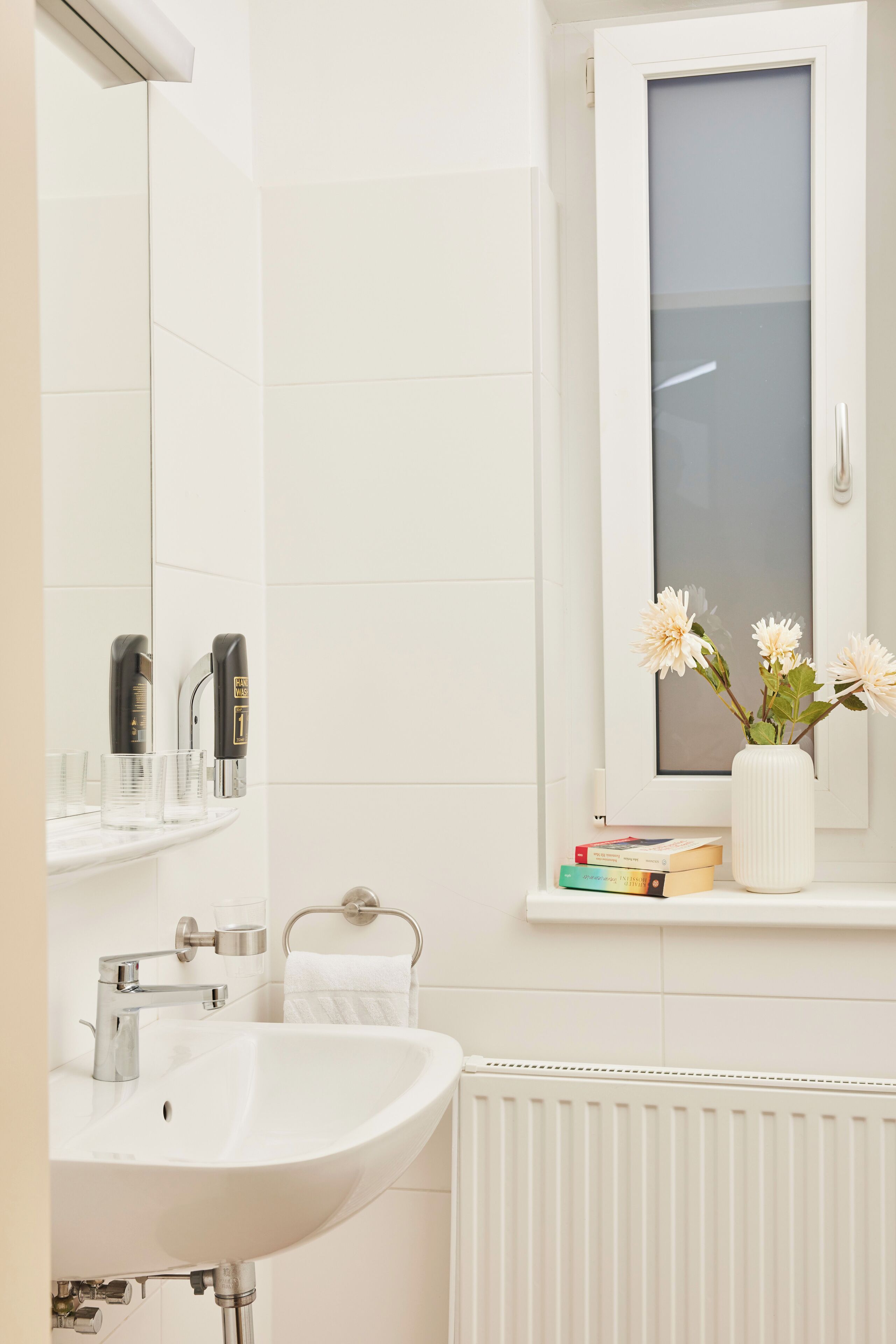 Un baño con lavamanos blanco, espejo y ventana. Hay artículos de aseo en el lavamanos, un perchero y un florero con flores en el alféizar.
