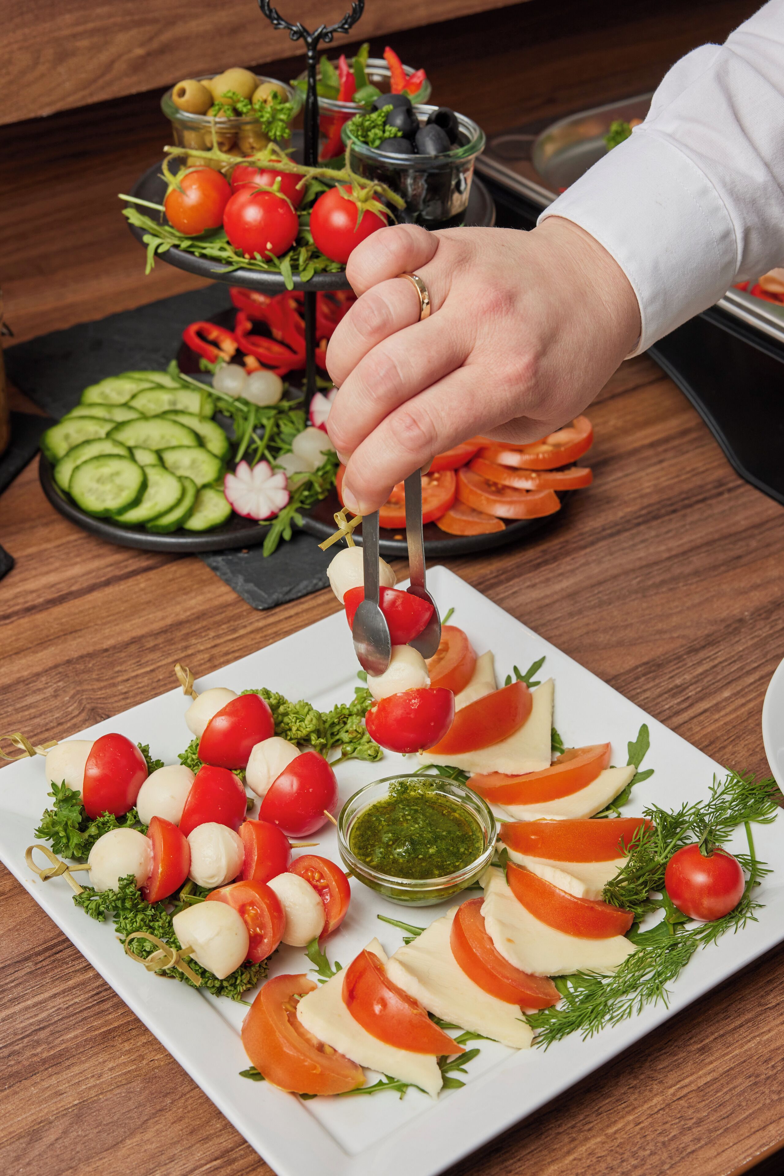 A plate of food with a hand holding a spoon, a bowl of olives, and a plate of sliced vegetables.