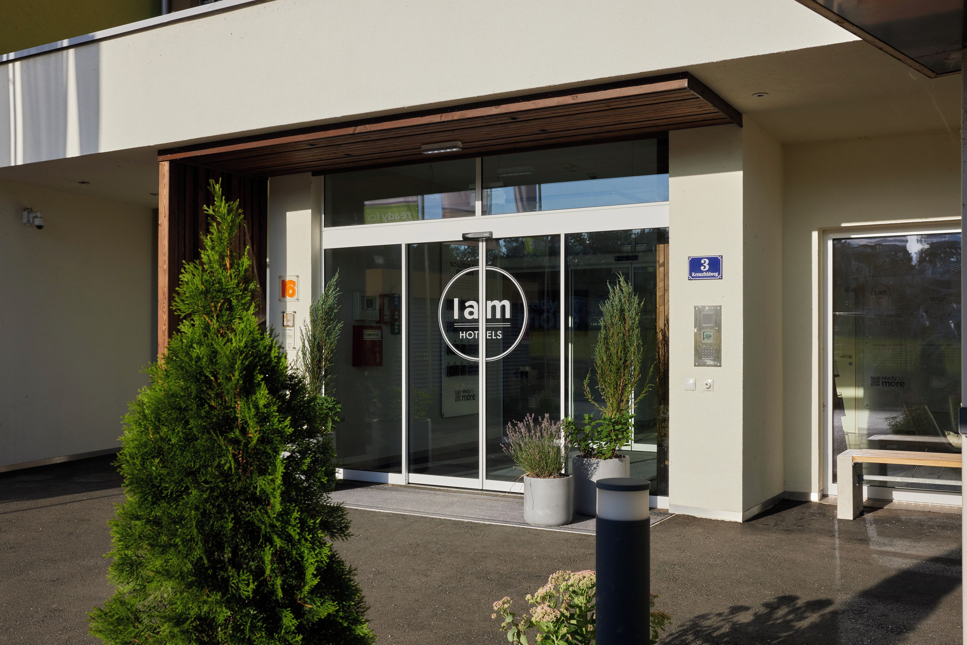 En hotelindgang med glaskonstruktion, et skilt der viser '3 Kreuzfeldweg', og en bænk foran.