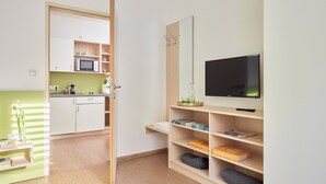 A modern, well-lit room with a wooden floor, a television, a bookshelf, and a kitchen area visible through an open door. - I AM Hotel Leoben (Leoben)