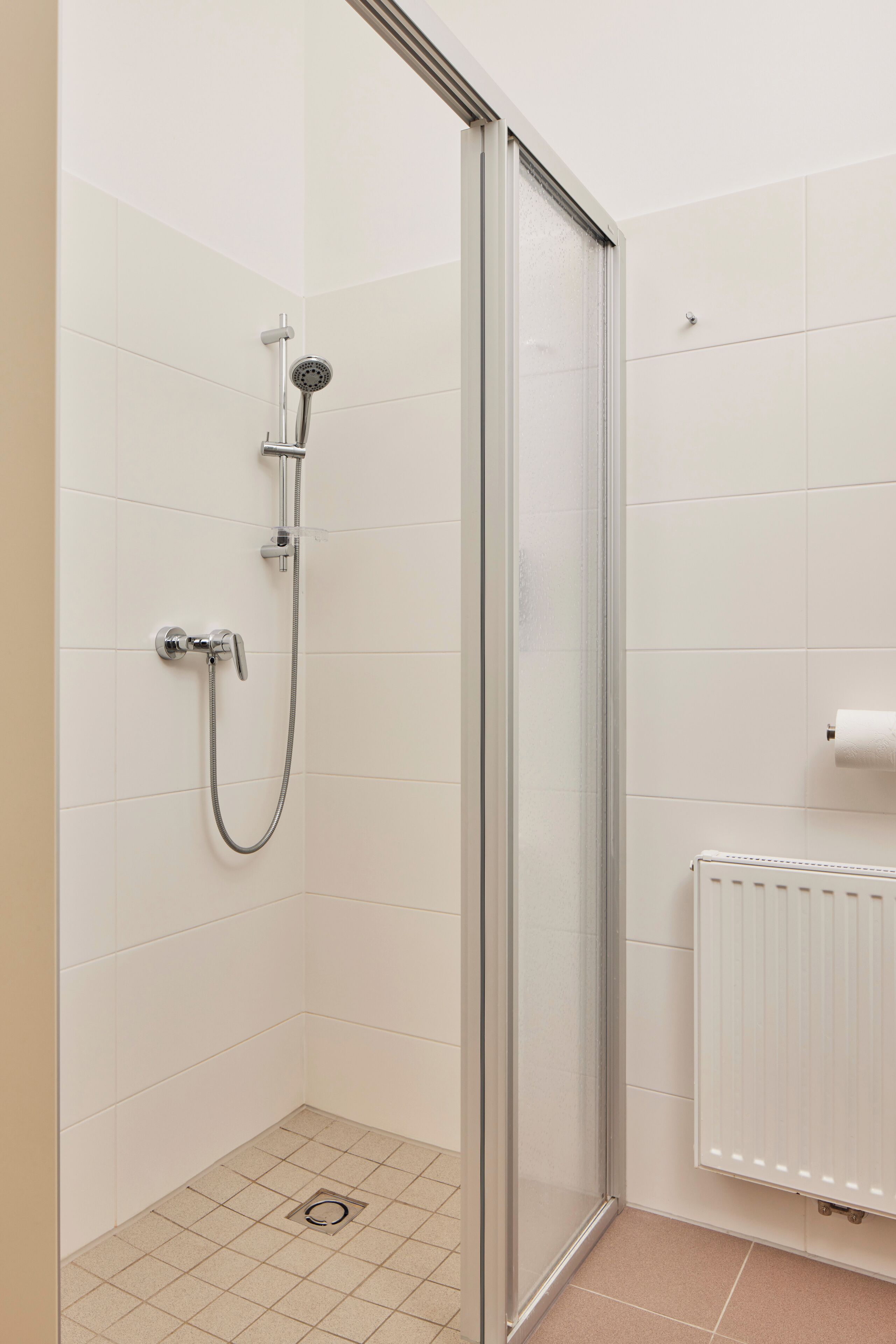 Eine Dusche mit Glastür, ein Handtuchspender und ein Heizkörper in einem gefliesten Badezimmer.