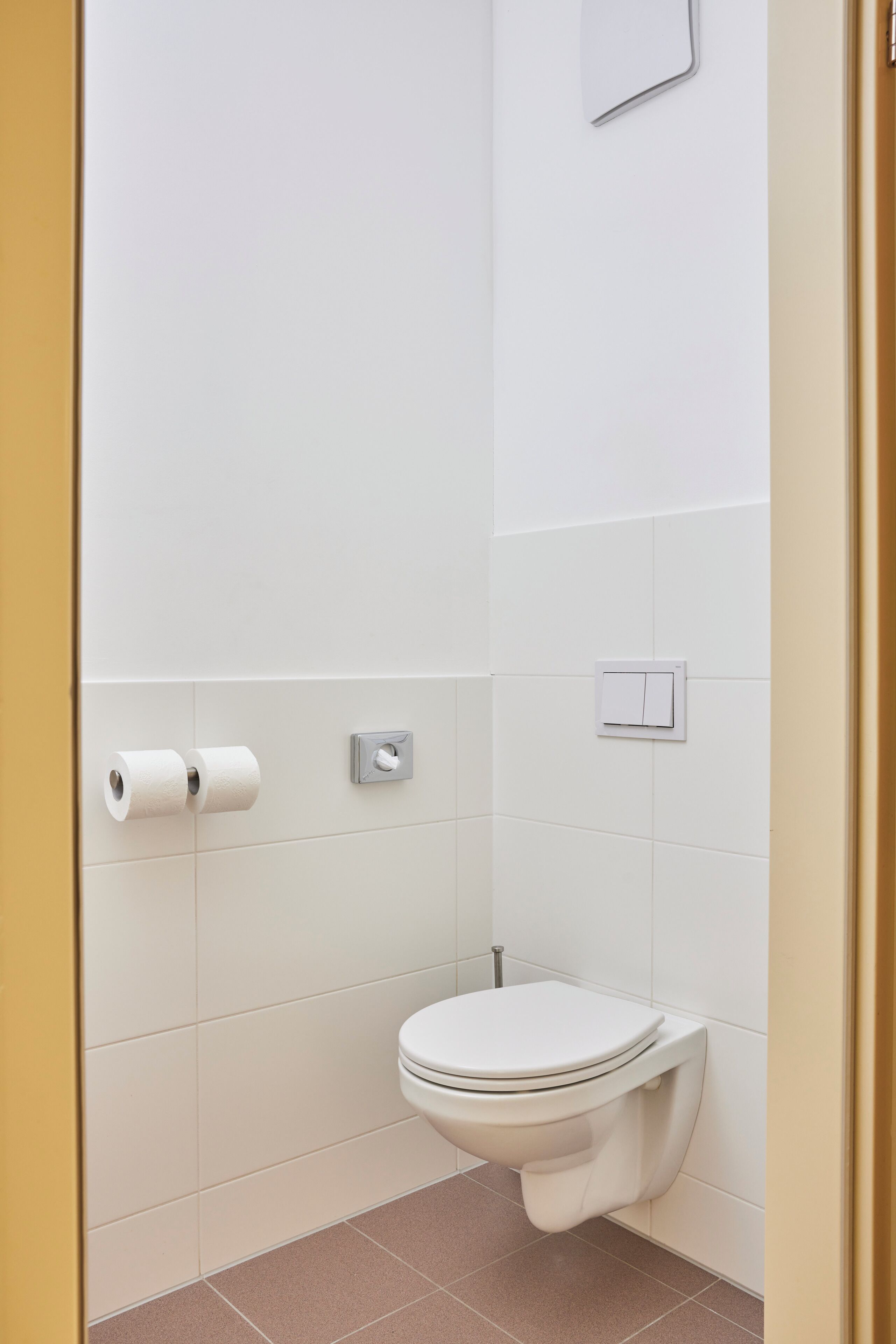 A small, clean bathroom with a toilet, two rolls of toilet paper, and a light switch on the wall.