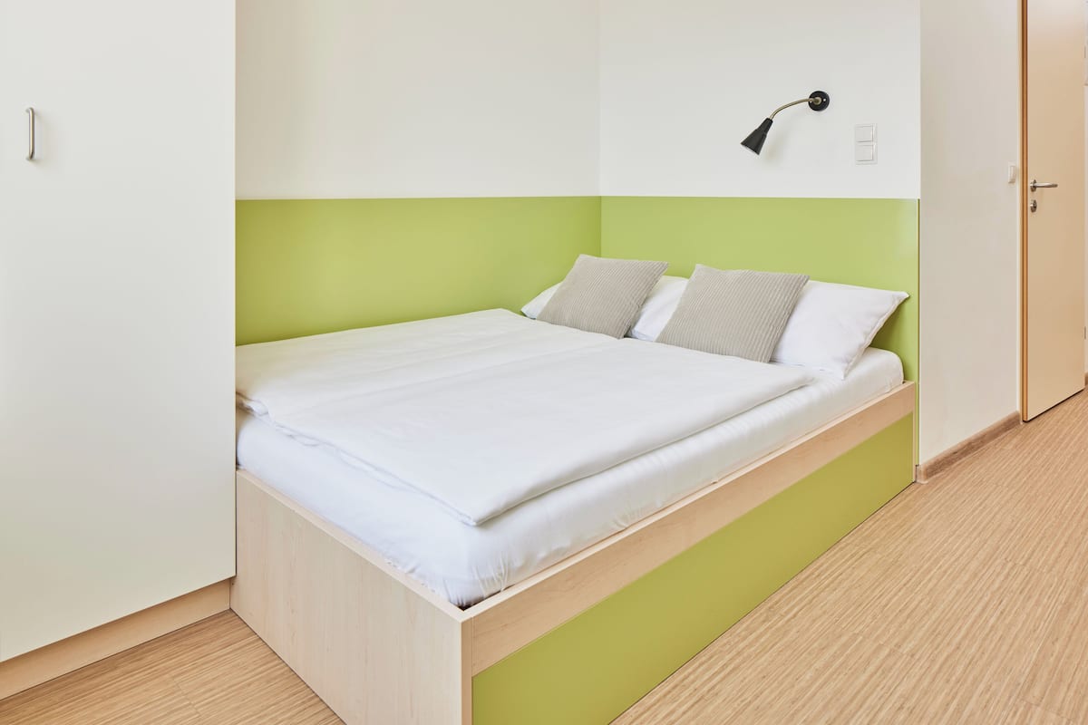 Una cama individual con estructura de madera y cabecero verde en una habitación con paredes blancas y suelo de madera.
