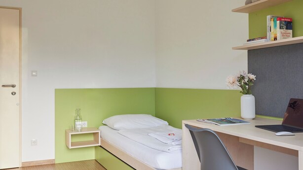 A bedroom with a bed, desk, chair, and a small shelf with books and a vase. - I AM HOTELS Leoben (Leoben)