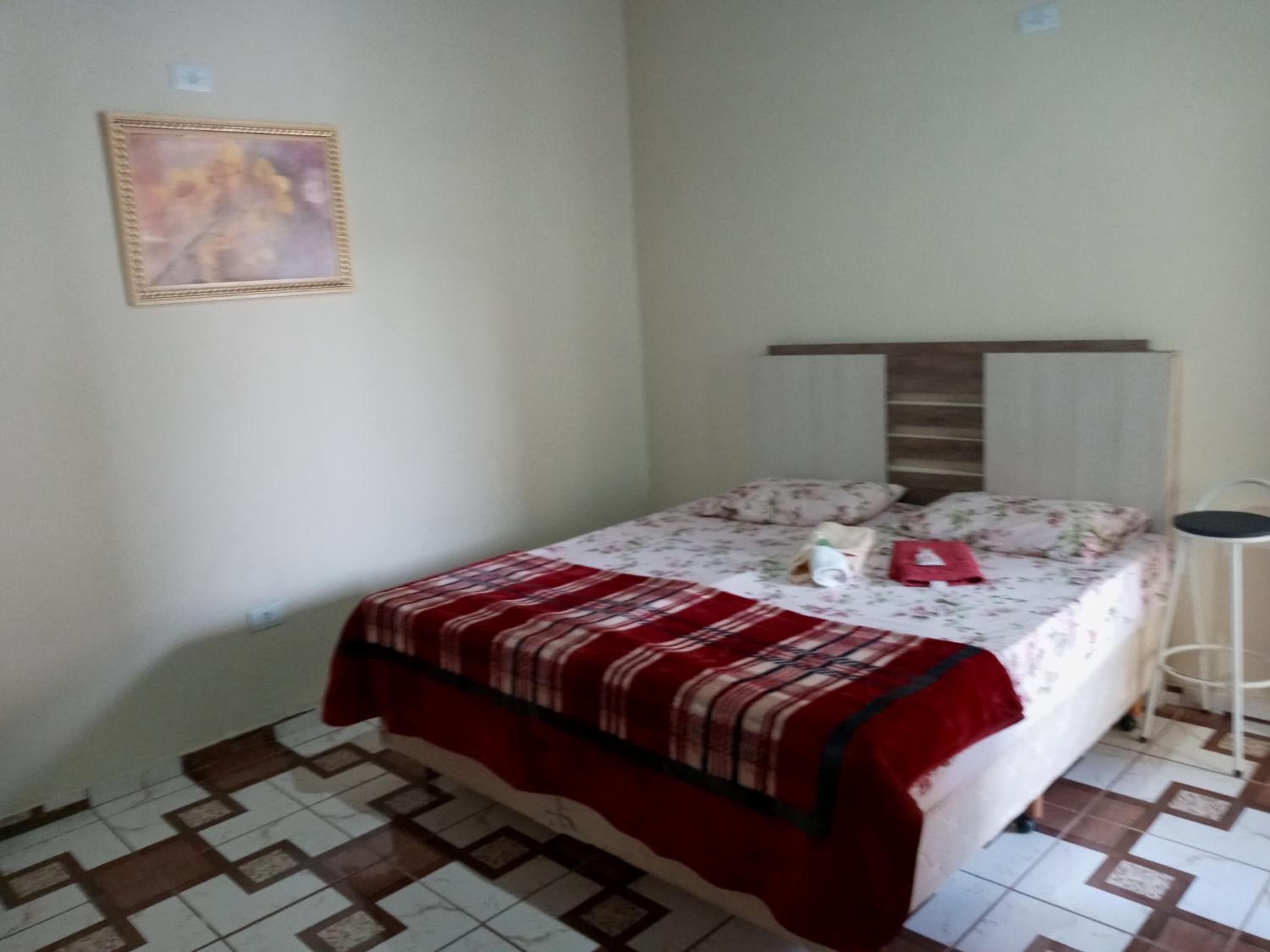 Una cama individual con colcha estampada y una manta roja, una mesita de noche y una lámpara de pie.