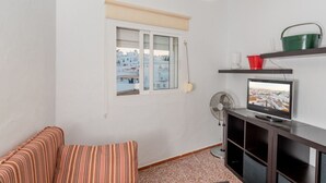 Ein kleines Wohnzimmer mit einer Couch, einem Fernseher, einem Ventilator und einem Fenster mit Blick auf Gebäude.