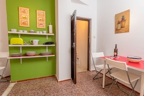 Una habitación con pared verde, una mesa blanca con sillas, mantel rojo y una puerta que da a otra habitación.