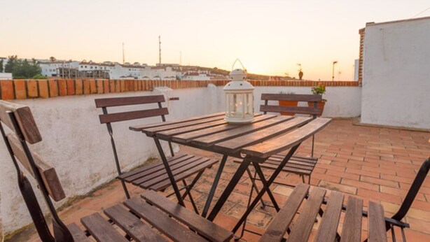 Eine Dachterrasse mit Holztisch und Bänken, einer Laterne und Blick auf die Stadt bei Sonnenuntergang.