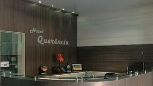 A hotel reception desk with a wooden panel wall, a glass counter, and a sign that reads "Hotel Querencia". - Hotel Querência (Pitanga)